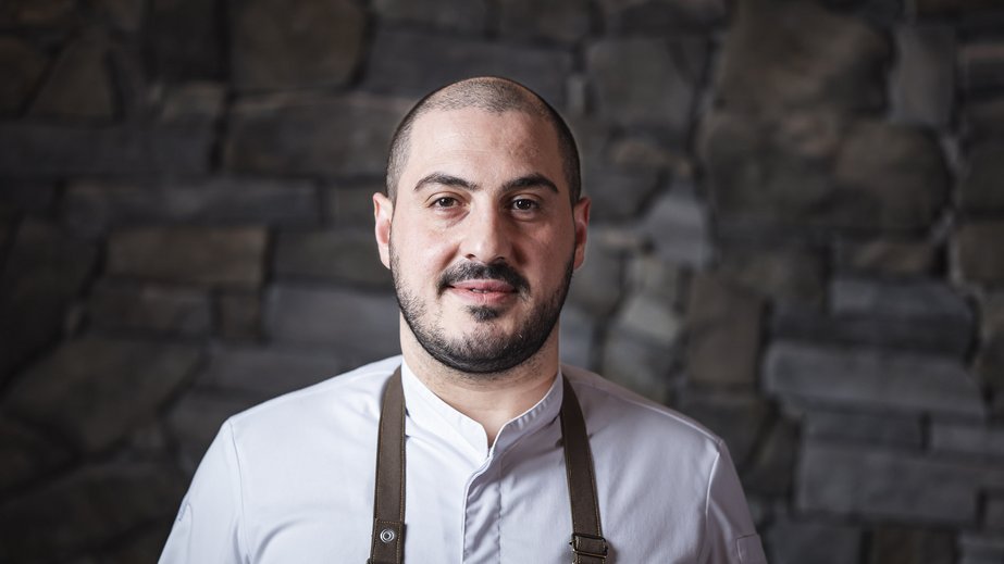 Il nostro hotel a Valdaora tra cielo e terra Ritratto di uno chef con giacca bianca e grembiule nero davanti a muro di pietra