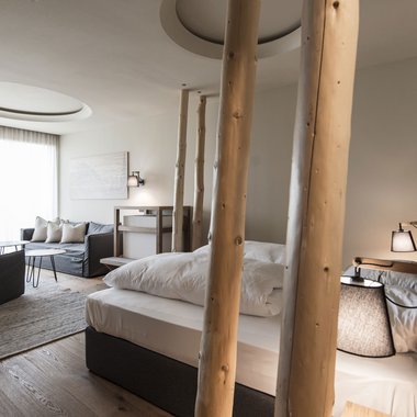 Benessere su tutte le sfaccettature dell’essere Camera d'albergo moderna con dettagli in legno, letto e zona soggiorno con divano e poltrone