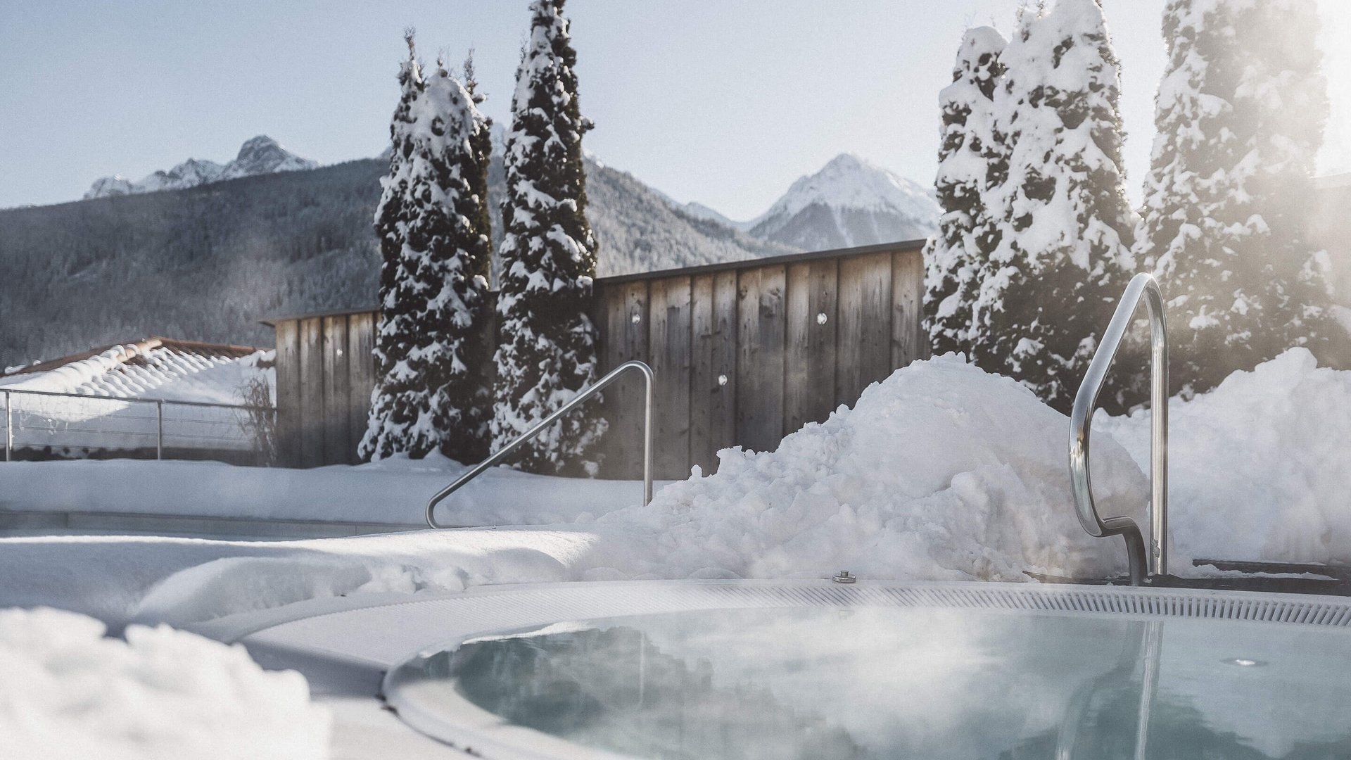 Il nostro hotel con infinity pool in Alto Adige: una meraviglia Vasca idromassaggio all'aperto con alberi e montagne innevate sullo sfondo.