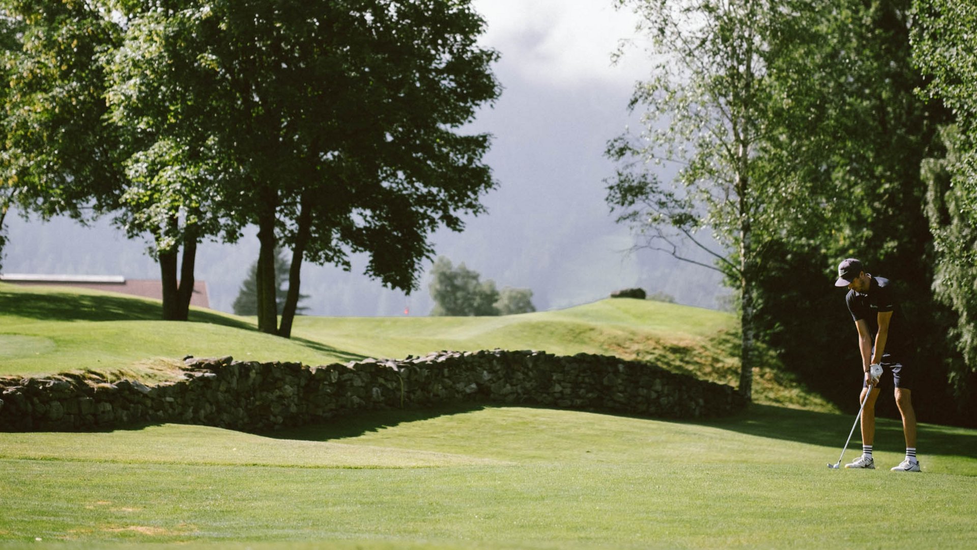 Golf Club Pustertal - Il tuo campo di golf a Plan de Corones Persona gioca a golf su un campo verde con alberi e montagne sullo sfondo