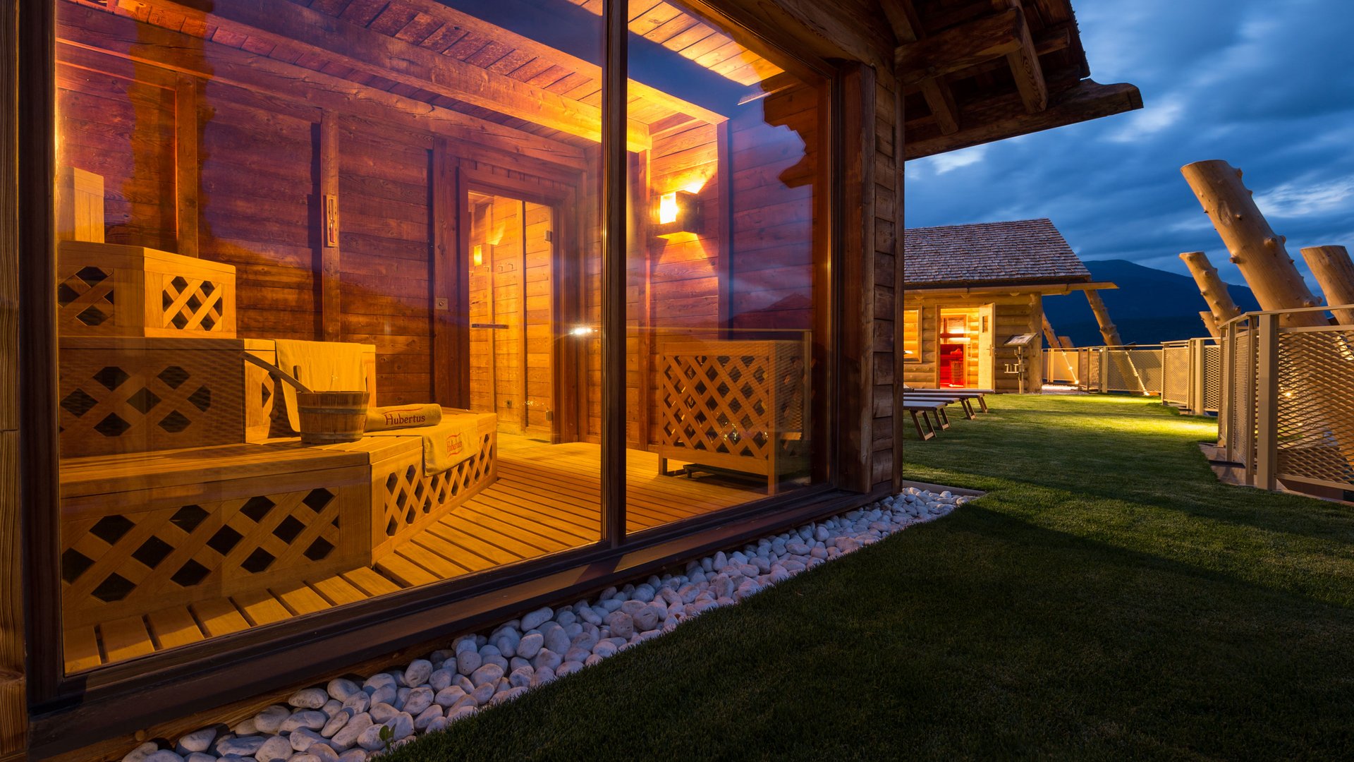 Unsere Sauna, den Kronplatz im Blickfeld Beleuchtete Holzsauna mit Glaswand und Außenbereich bei Abenddämmerung