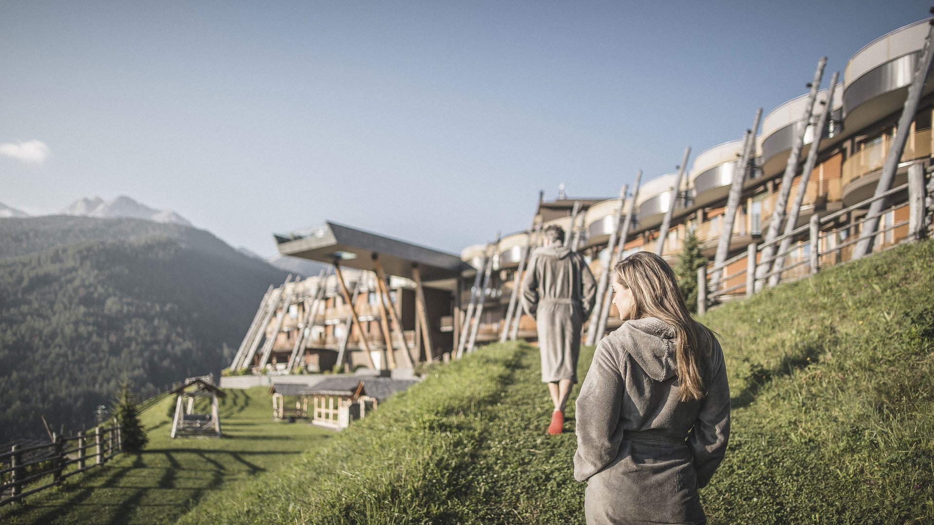 Erholung auf allen Ebenen des Seins Zwei Personen in Bademänteln gehen an einem Berghotel im Grünen entlang
