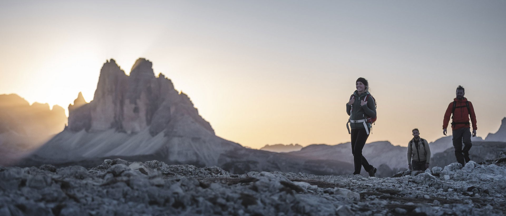 Ein Wanderurlaub bei den Spezialisten Drei Wanderer auf felsigem Pfad mit Berg im Sonnenuntergang