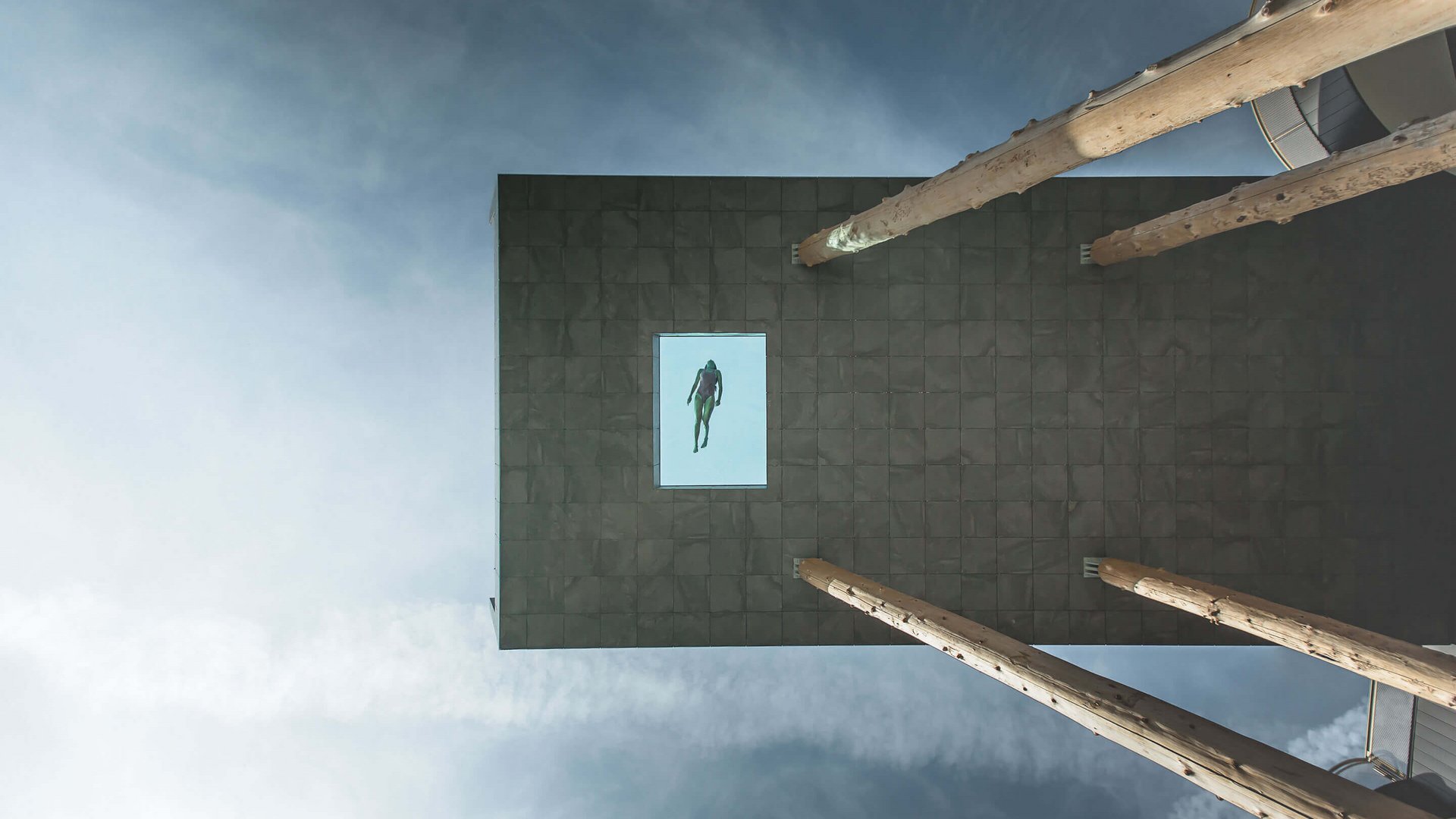 Unser Hotelgutschein: Südtirol im Geschenkformat Schwimmer schwimmt in einem Glasbecken auf einer hohen Plattform vor bewölktem Himmel