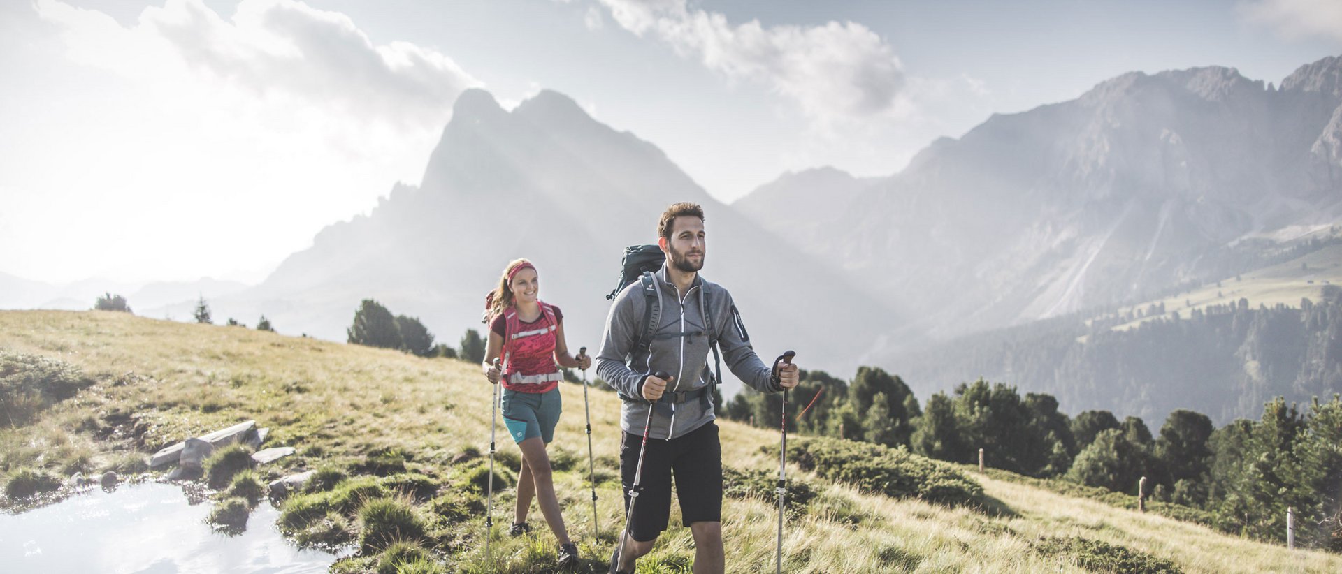 Ein Wanderurlaub bei den Spezialisten Zwei Wanderer mit Rucksäcken und Stöcken auf Bergwiese bei Sonnenschein