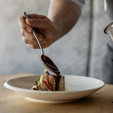 Sommerurlaub am Kronplatz: unvergesslich Koch gießt Soße über ein kleines Stück Fleisch auf einem weißen Teller