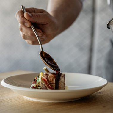 Bruneck: Shopping und mehr Koch gießt Soße über ein kleines Stück Fleisch auf einem weißen Teller